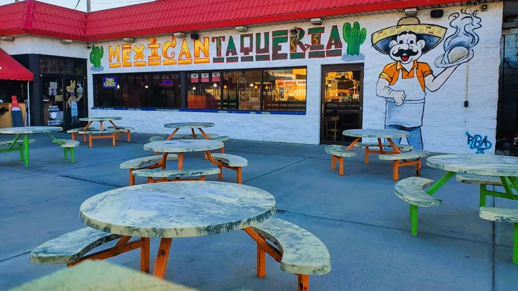 Colorful Mexican taqueria with mural, sign, and empty outdoor picnic tables.