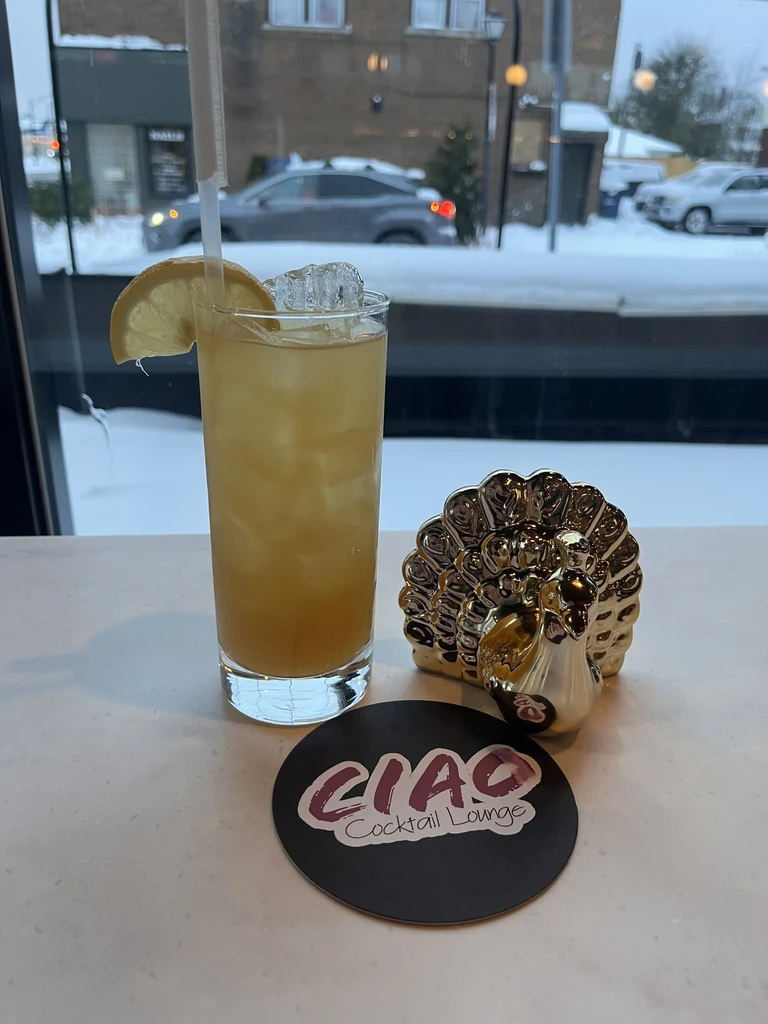 Amber cocktail, peacock figurine, and 'CIAO' coaster on a snowy day.