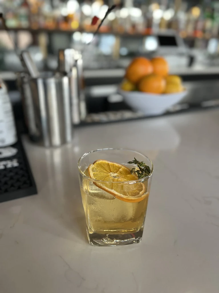 Light amber cocktail in short glass with dried orange and herb.
