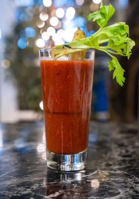Reddish-brown drink in glass with celery, pickle, on dark marble.