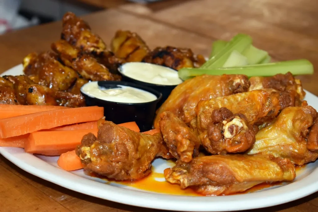 Close-up of saucy chicken wings, white dip, celery, and carrots.