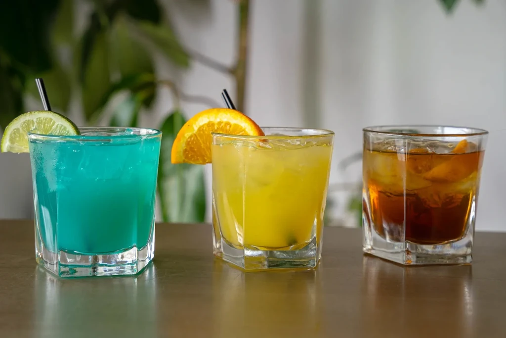 Row of three garnished blue, yellow, amber cocktails in faceted glasses.