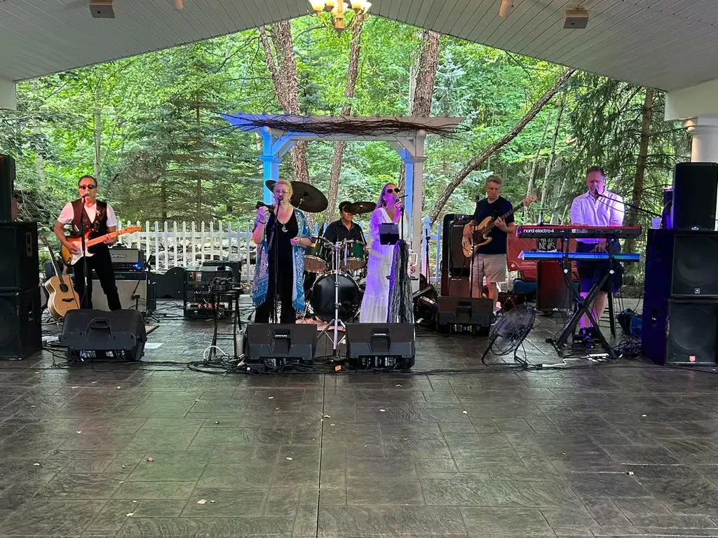Five musicians perform on a covered outdoor stage with trees.