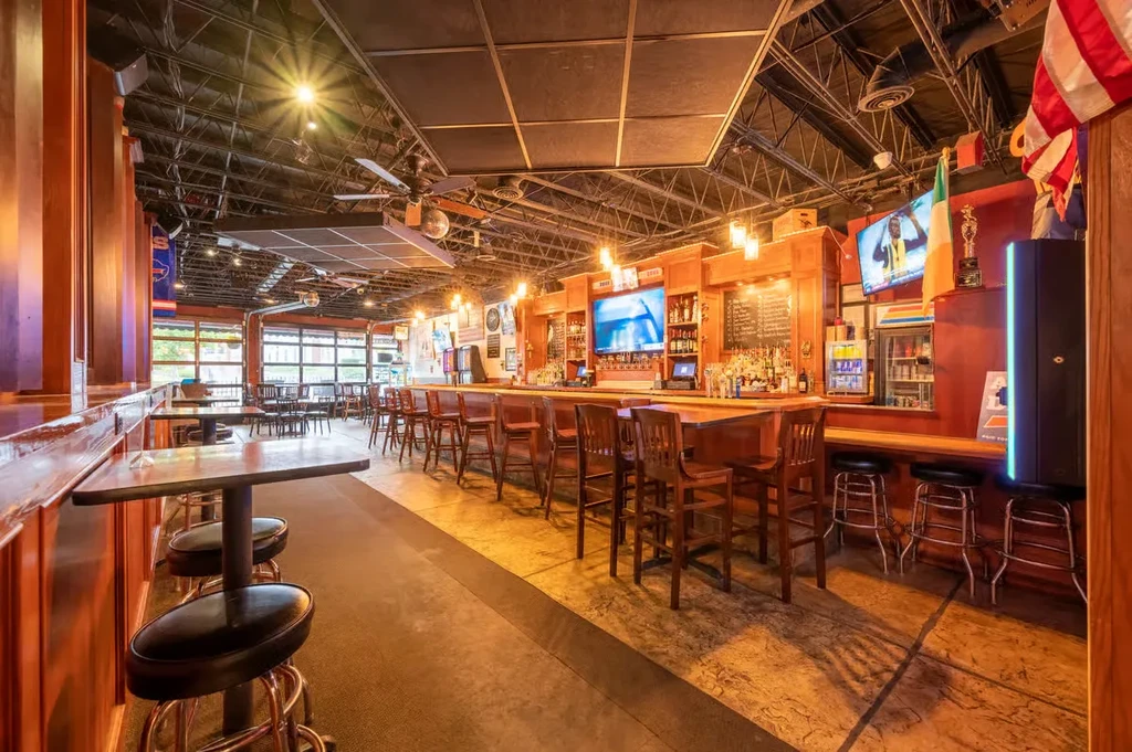 Empty bar interior with wooden counter, bottles, TVs, industrial ceiling.