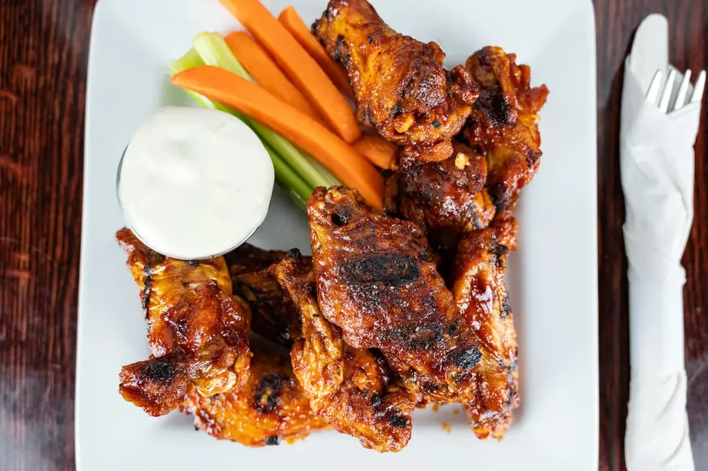 Glazed chicken wings, dipping sauce, carrot and celery sticks on white plate.