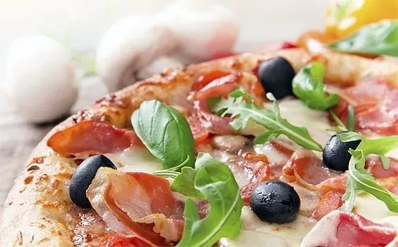 Close-up of pizza with prosciutto, black olives, basil, arugula, and cheese.