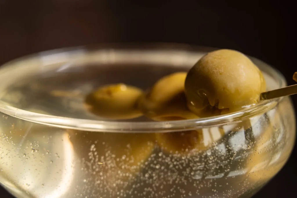 Martini glass with green olives and a pick on dark background.