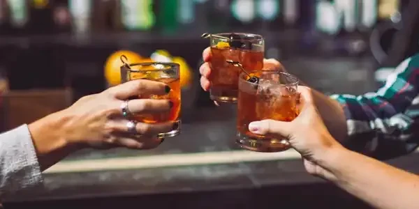 Three hands toast amber drinks with ice and fruit garnishes.