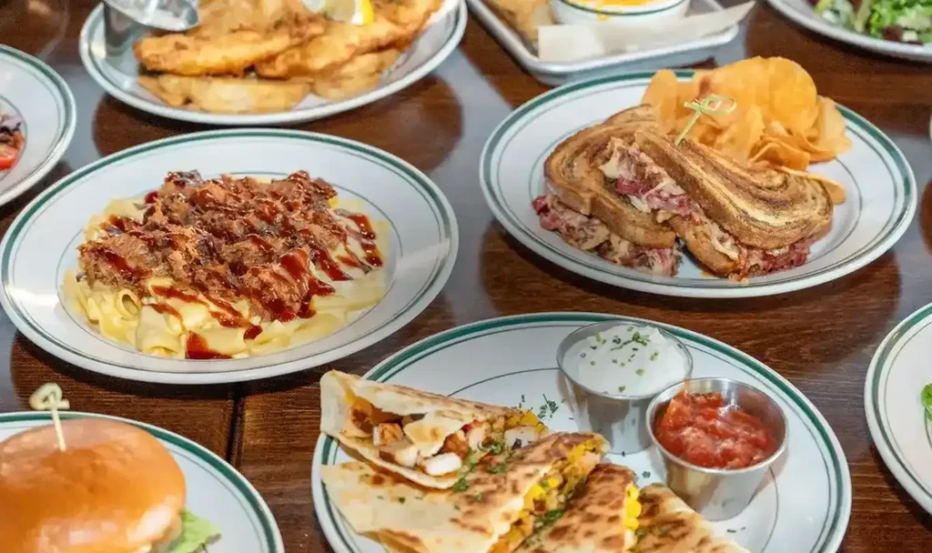 Overhead of quesadillas, pasta, Reuben, and more on a wooden table.