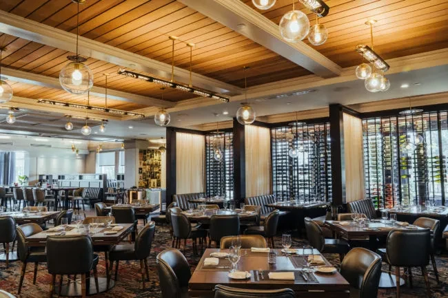 Warmly lit upscale dining room with set tables and floor-to-ceiling wine racks.
