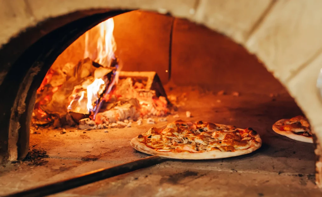 Golden pizza cooking in a rustic wood-fired oven with flames.
