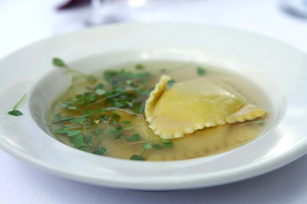 Single golden ravioli with crimped edges in clear broth with herbs.