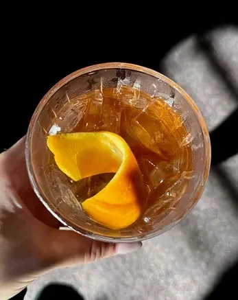 Top-down view of amber drink with ice, orange peel, and hand.