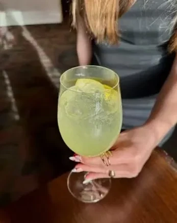 Hand holding a green drink with ice and citrus in a stemmed glass.
