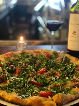 Gourmet pizza with arugula, tomatoes, sausage, wine, and candlelit background.