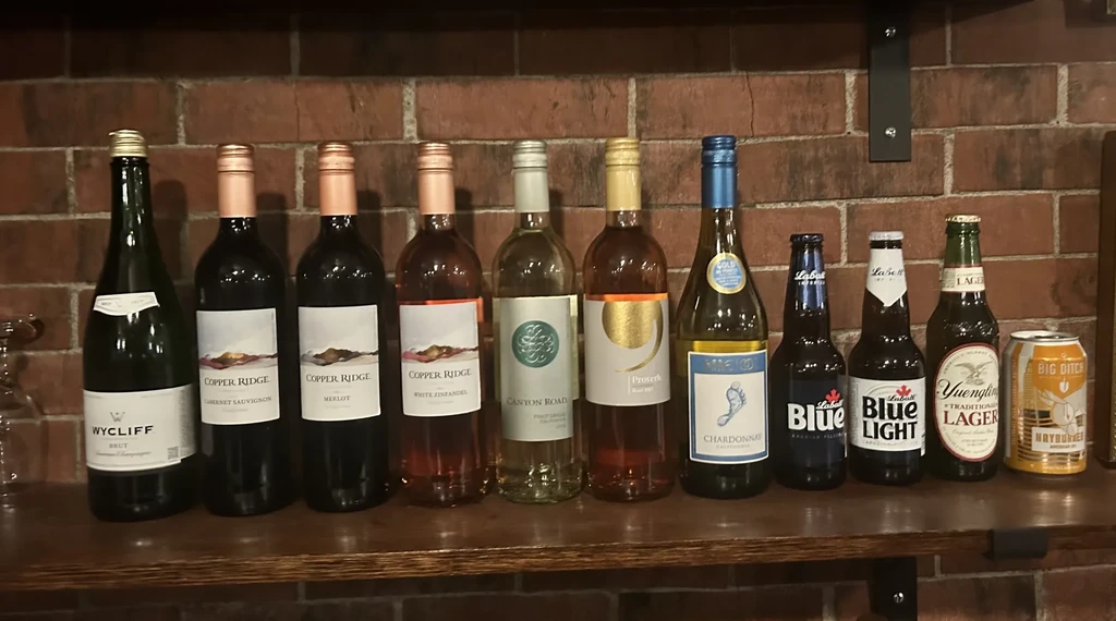 Eleven wine and beer bottles and a can on a wooden shelf.