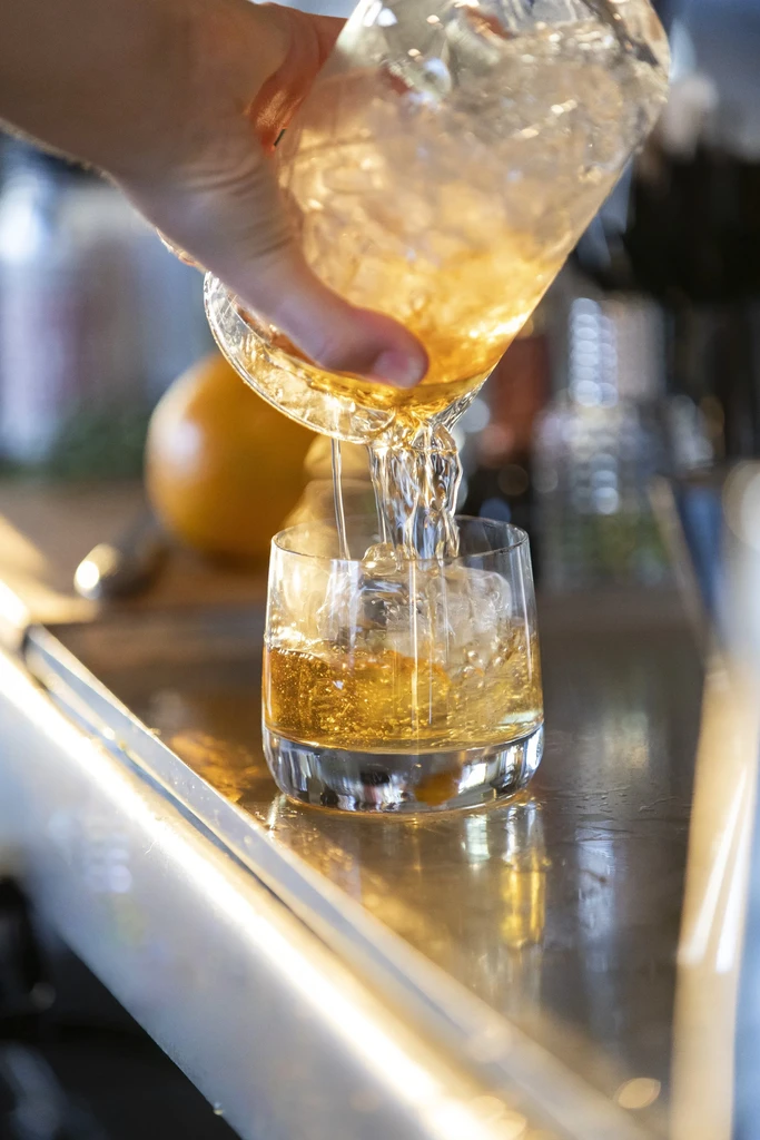 Hand pouring golden-amber drink into ice-filled lowball glass at a bar.