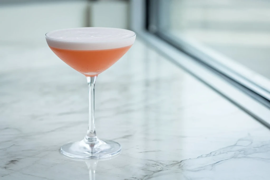 Two-layer cocktail in coupe glass: white foam, pink liquid, flecks.