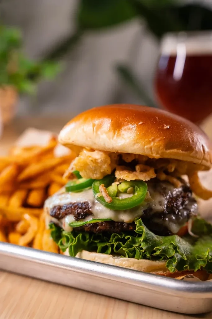 Gourmet burger with cheese, jalapeños, fries, and beer on a tray.