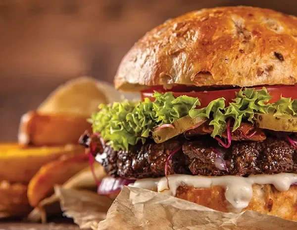 Close-up gourmet burger, partially wrapped in paper, with potato wedges.