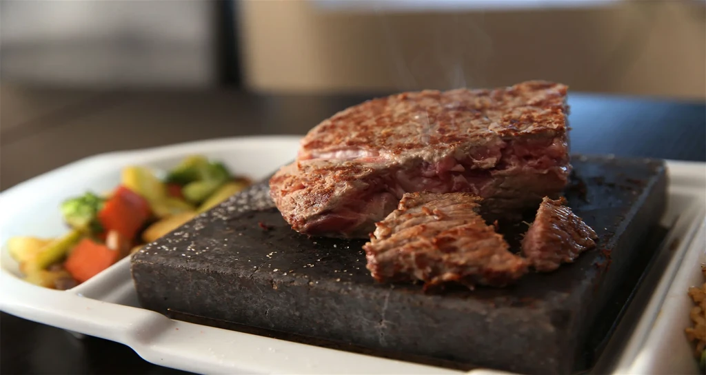 Thick-cut medium-rare steak on a steaming hot stone with colorful vegetables.