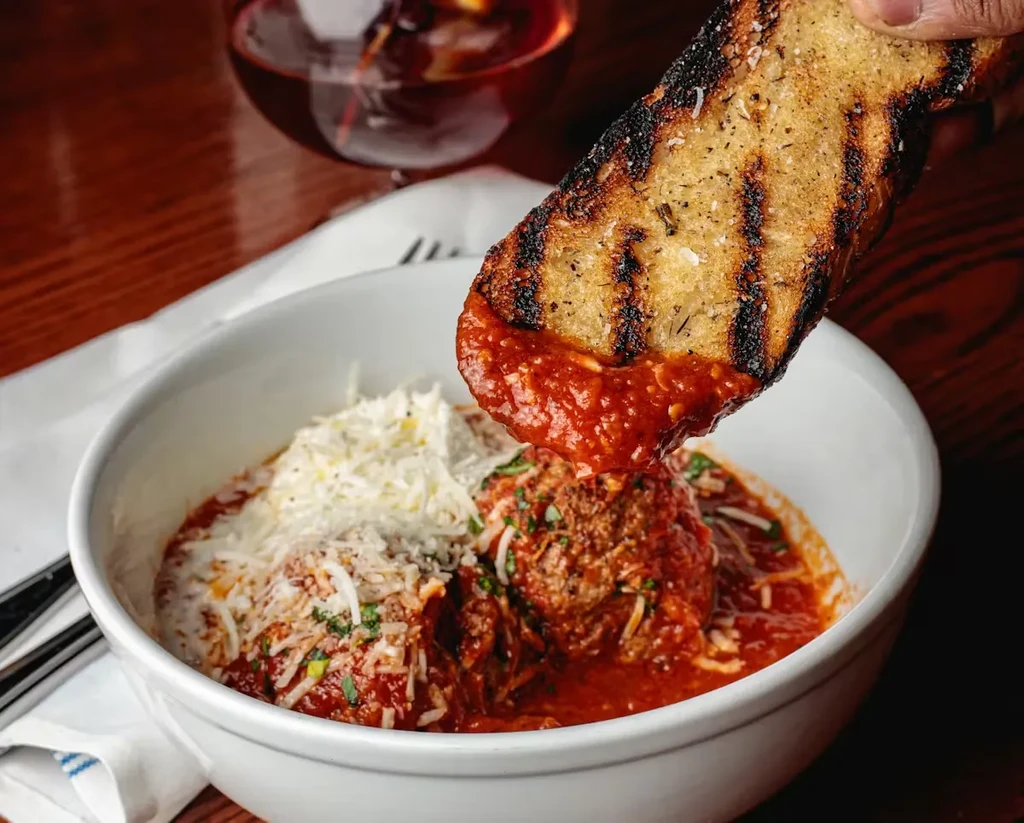 Hand dips grilled bread into meatballs with cheese and red sauce.