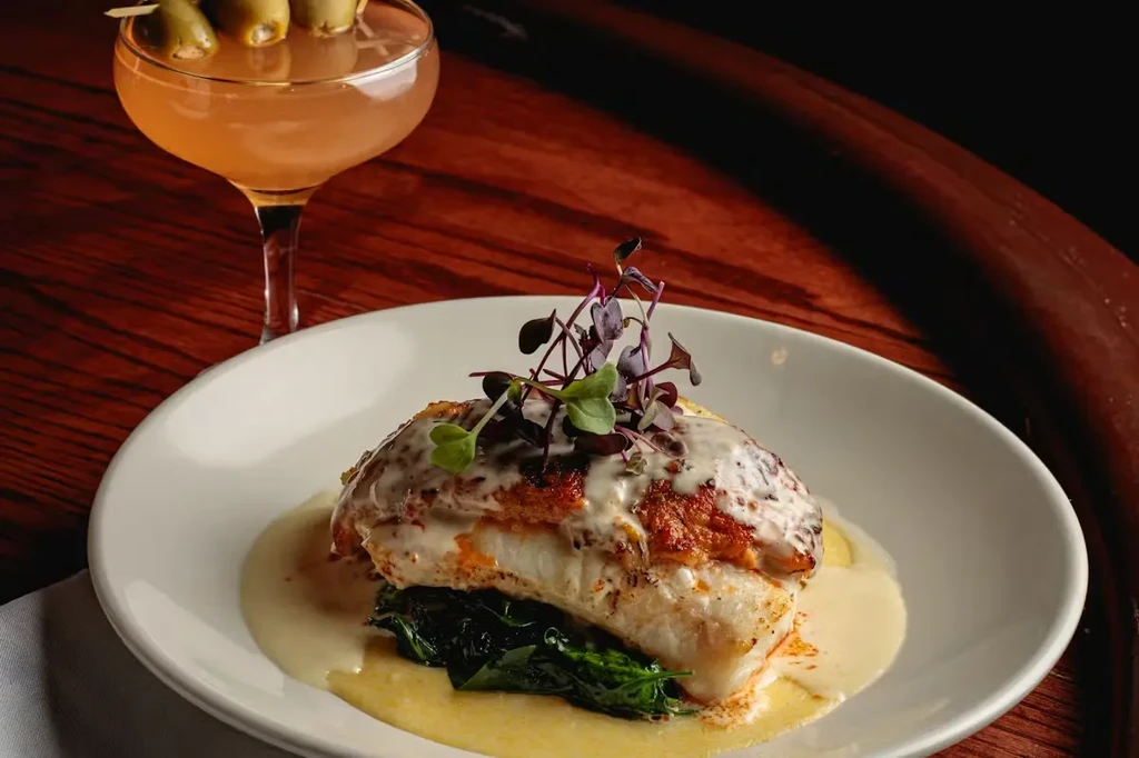 Pan-seared fish, leafy greens, creamy sauce, and olive cocktail on dark wood.