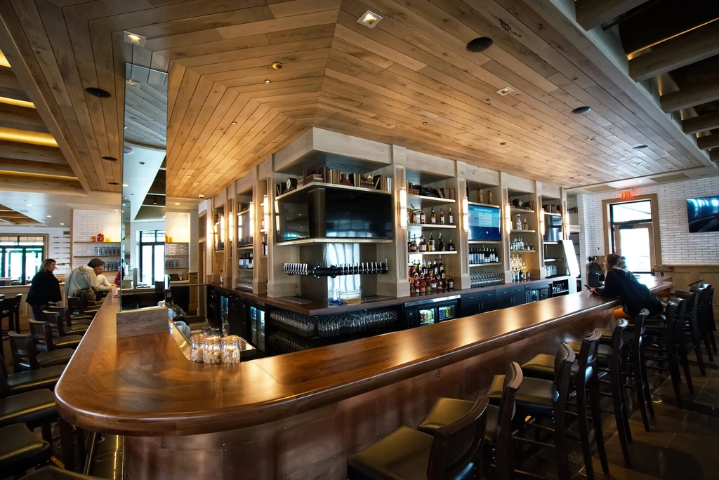 Modern dark wood bar with liquor shelves, patrons, and patterned ceiling.