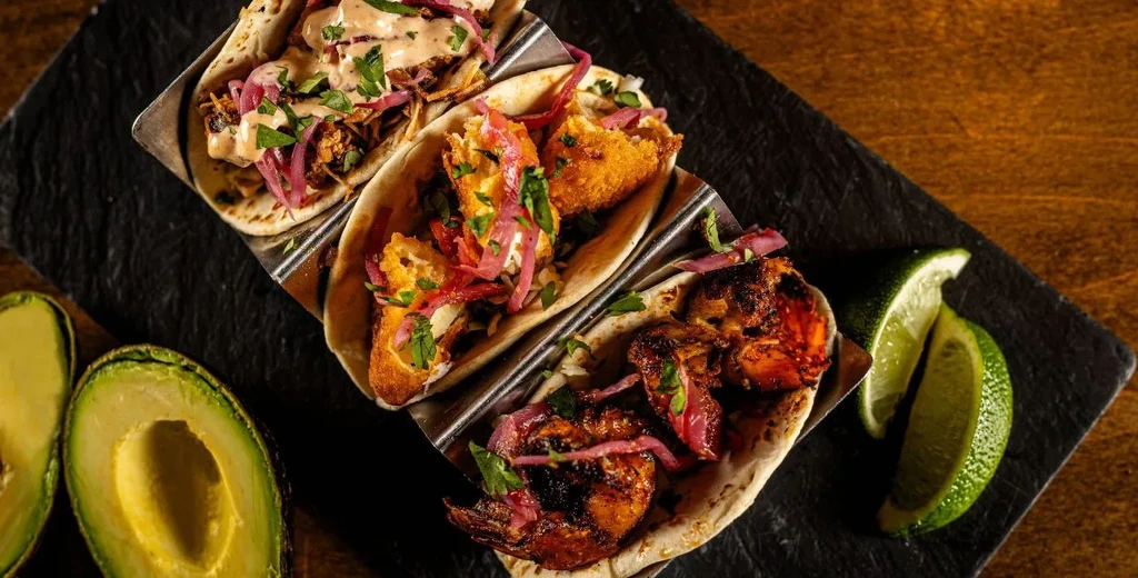 Three gourmet tacos (meat, crispy, shrimp) with avocado and lime.