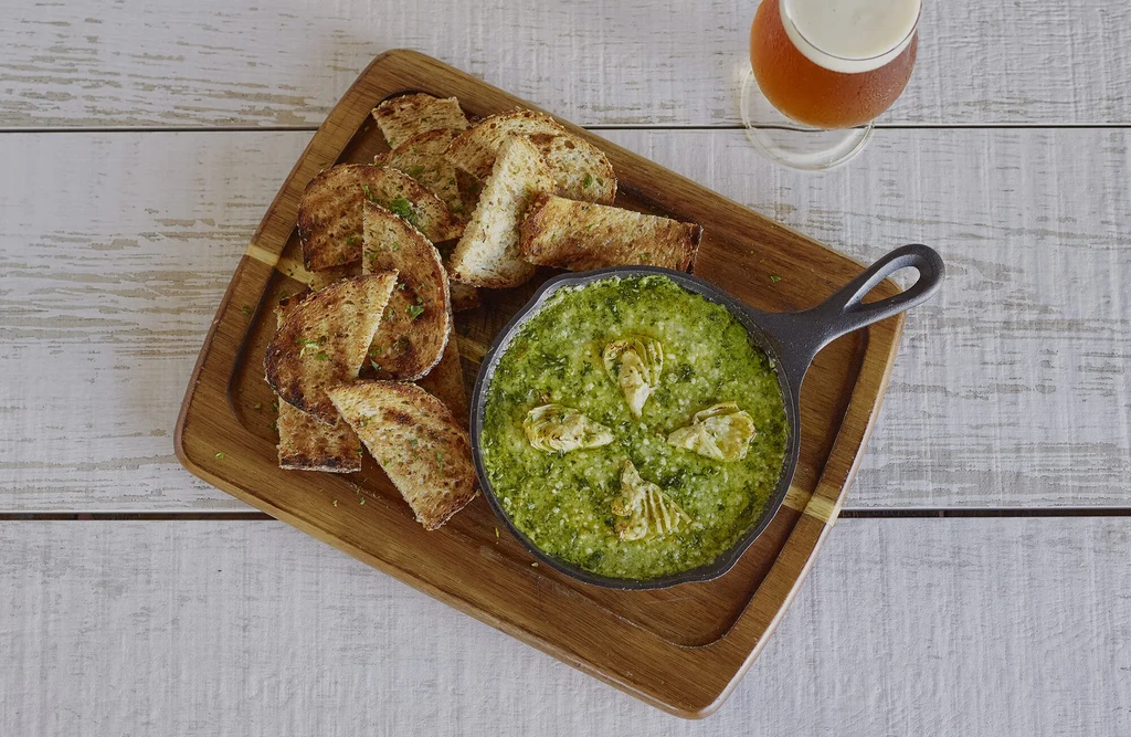 Overhead: Rustic board with artichoke dip, crostini, and amber beer.