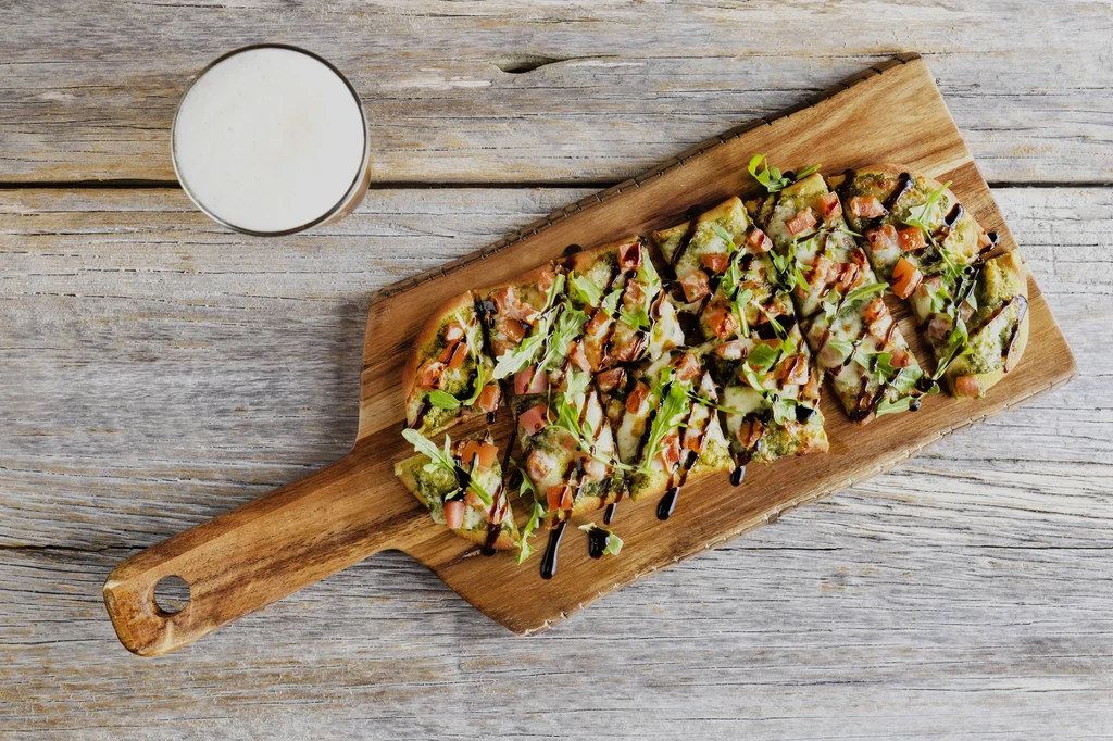 Flatbread with pesto, cheese, tomatoes, arugula, and beer on rustic table.