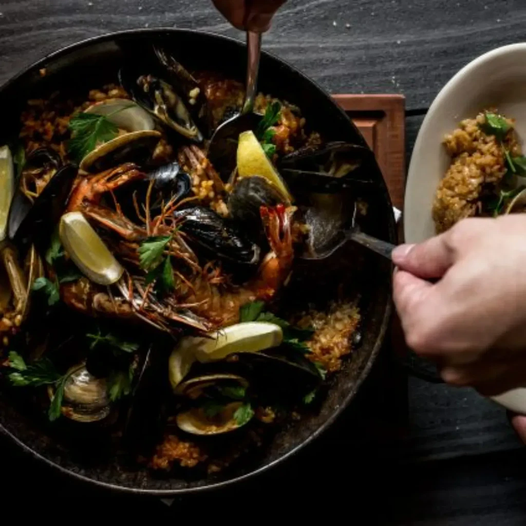 Close-up of hands serving golden-brown seafood paella from a dark pan.