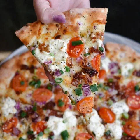 Hand holds a pizza slice topped with cheese, tomatoes, bacon, onion, and chives.