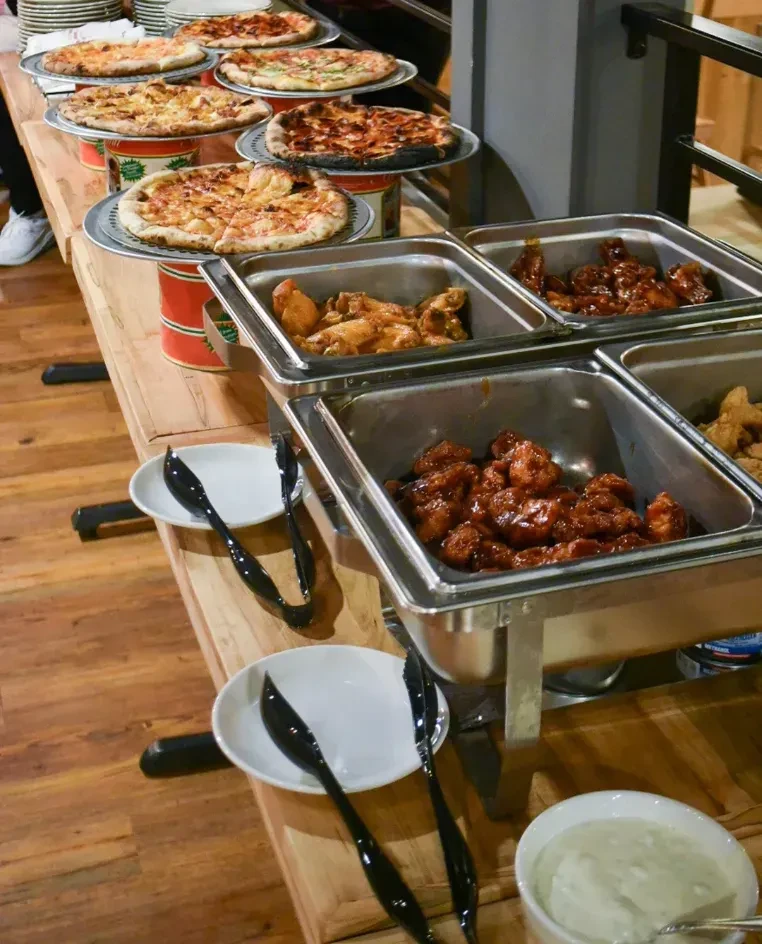 A buffet table showcases pizzas, chicken dishes, and various sauces.
