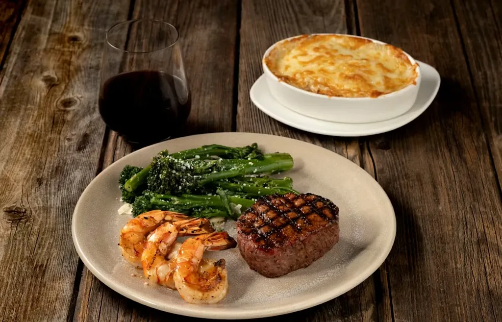 Grilled steak, shrimp, green vegetables, wine, and gratin on wooden table.