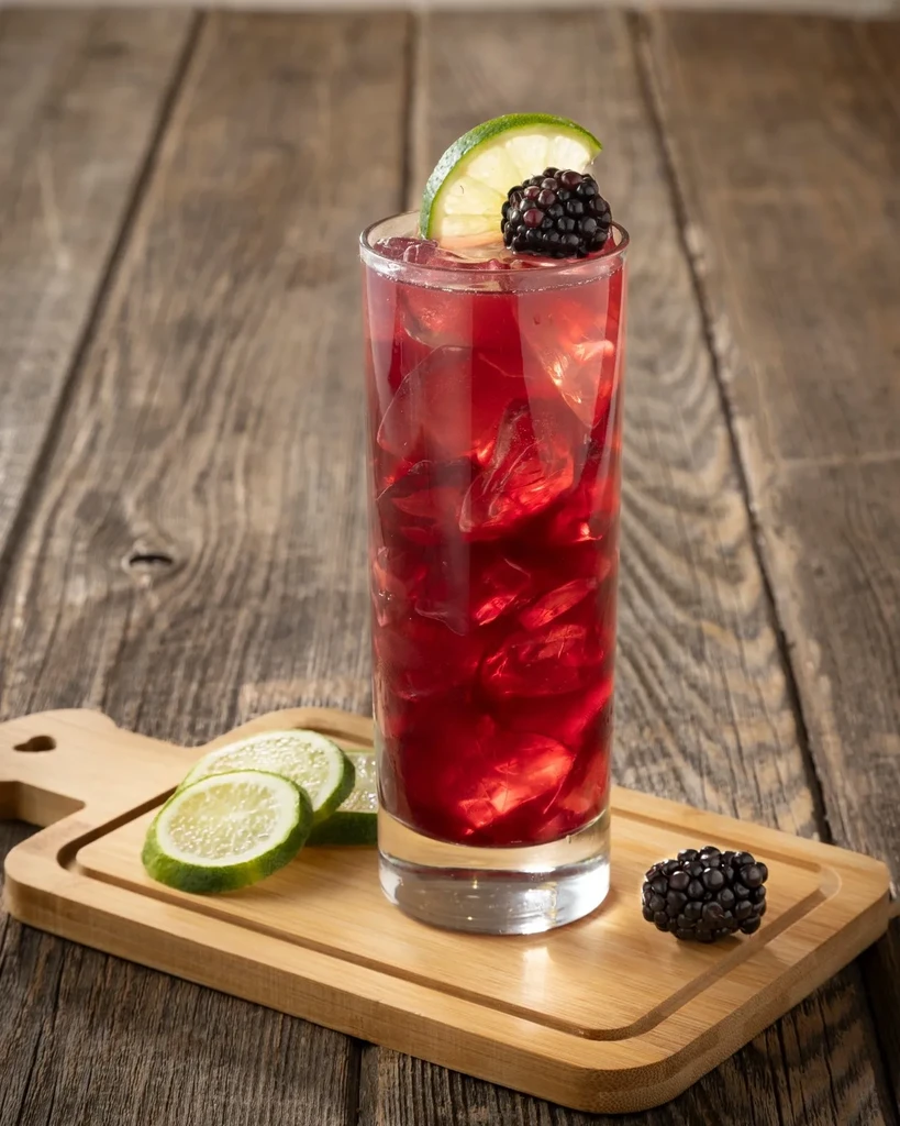 Red drink with ice, lime, and blackberry on a wooden board.