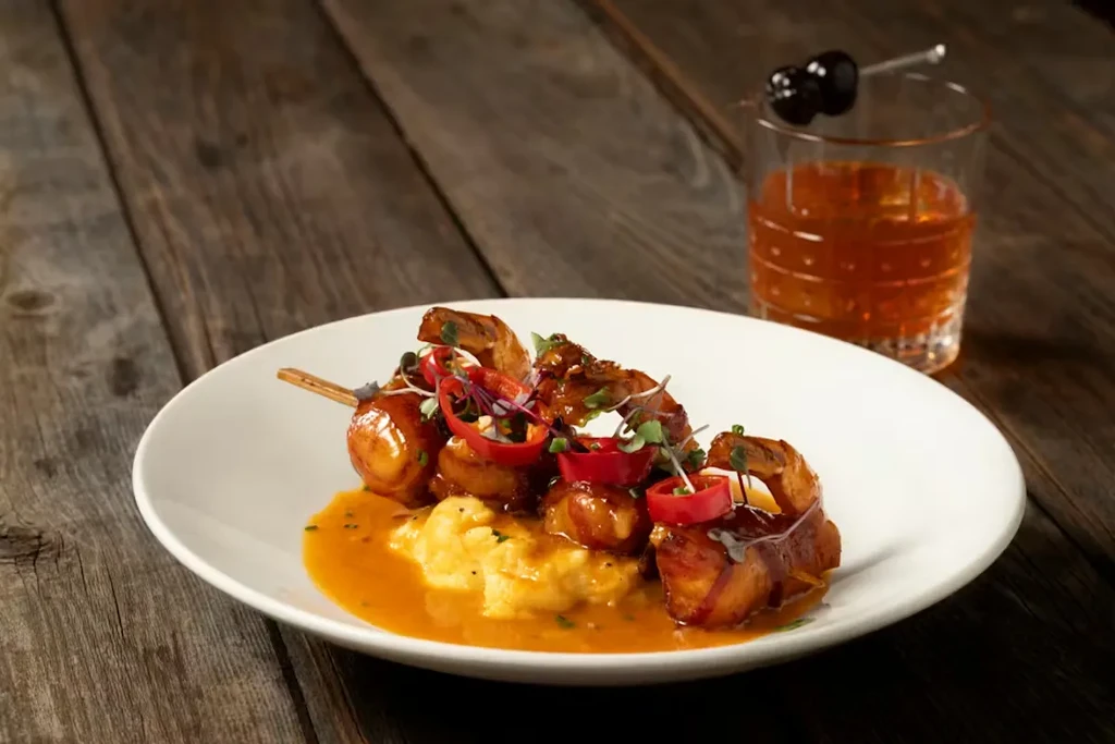 Gourmet bacon-wrapped seafood skewers with sauce, greens, and a cherry cocktail.