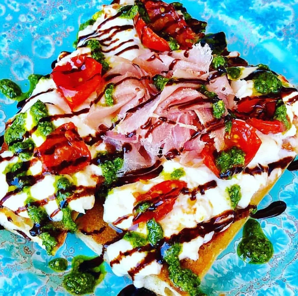 Bruschetta with mozzarella, prosciutto, tomatoes, pesto, and balsamic on a blue plate.