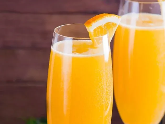 Two flutes with orange mimosas, one garnished with an orange slice on wood.