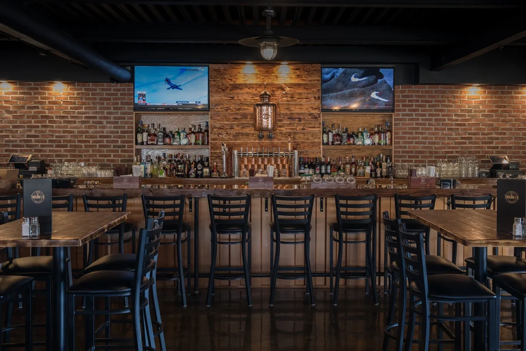 Empty industrial bar with a well-stocked counter, brick wall, and TVs.