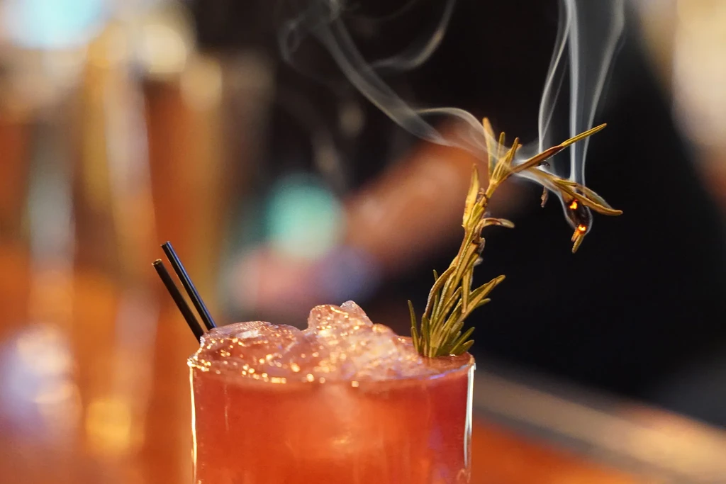 Red-orange cocktail with crushed ice, two straws, and smoking rosemary garnish.