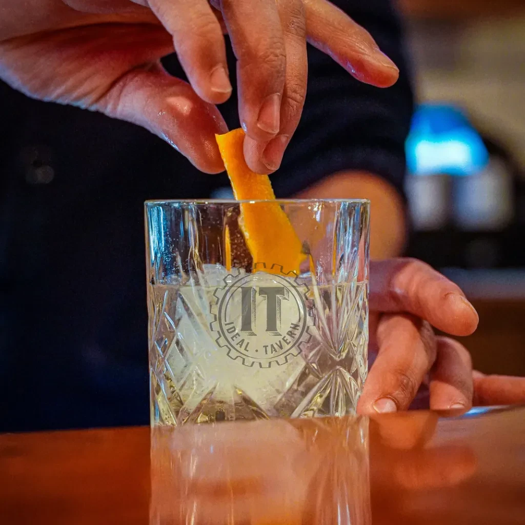 A person garnishes an 'Ideal Tavern' cocktail with an orange peel.