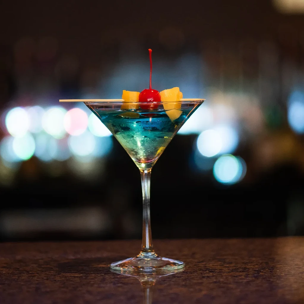 Shimmering blue cocktail in martini glass with cherry and pineapple.
