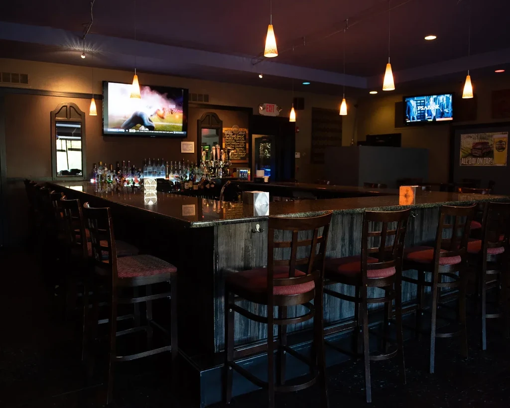 Empty, dimly lit bar with counter, red stools, liquor bottles, TVs.