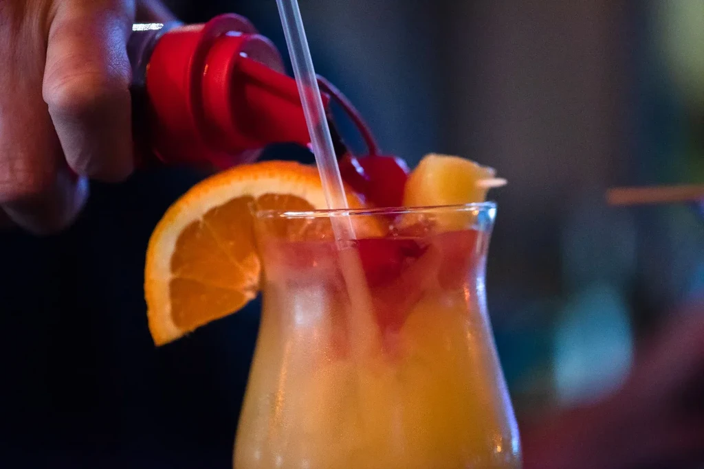 Hand pouring into a vibrant cocktail with orange, cherry, and pineapple garnishes.