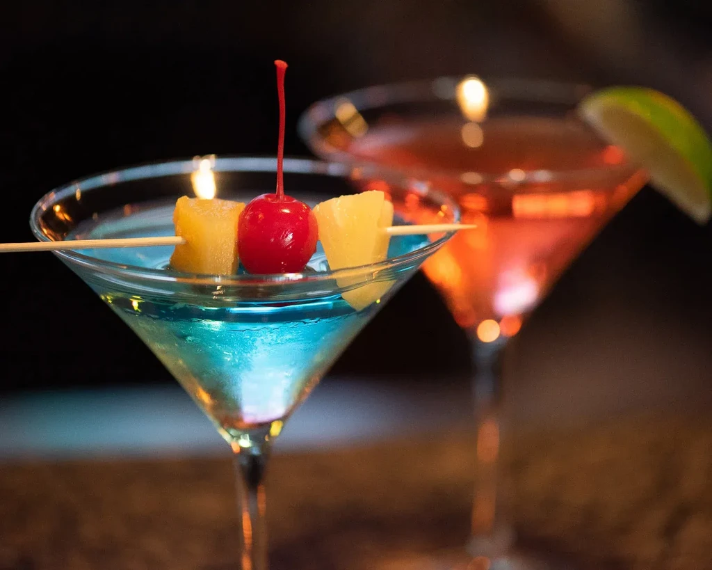 Brilliant blue cocktail with pineapple and cherry, blurred orange cocktail behind.