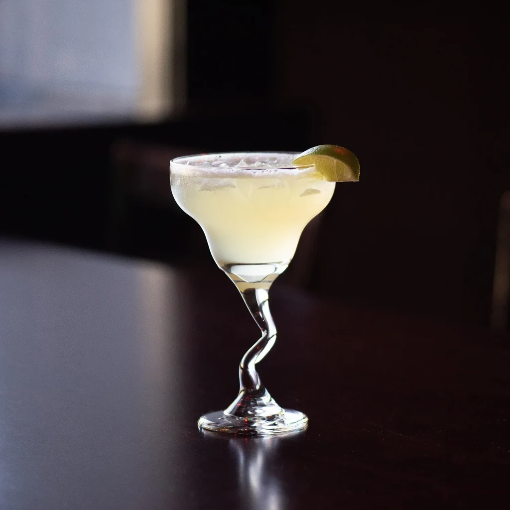 Cold pale yellow cocktail with lime in a wavy-stemmed glass.