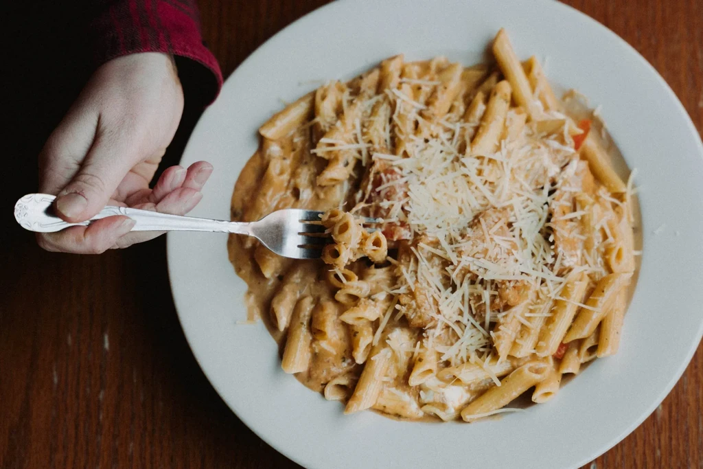 Close-up: Penne pasta with creamy sauce, cheese, hand holding fork above.