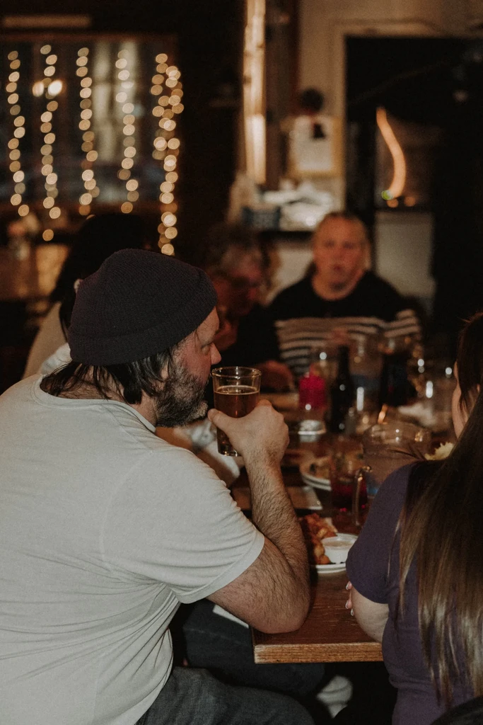Man in beanie drinking beer at a cozy, dimly lit restaurant.