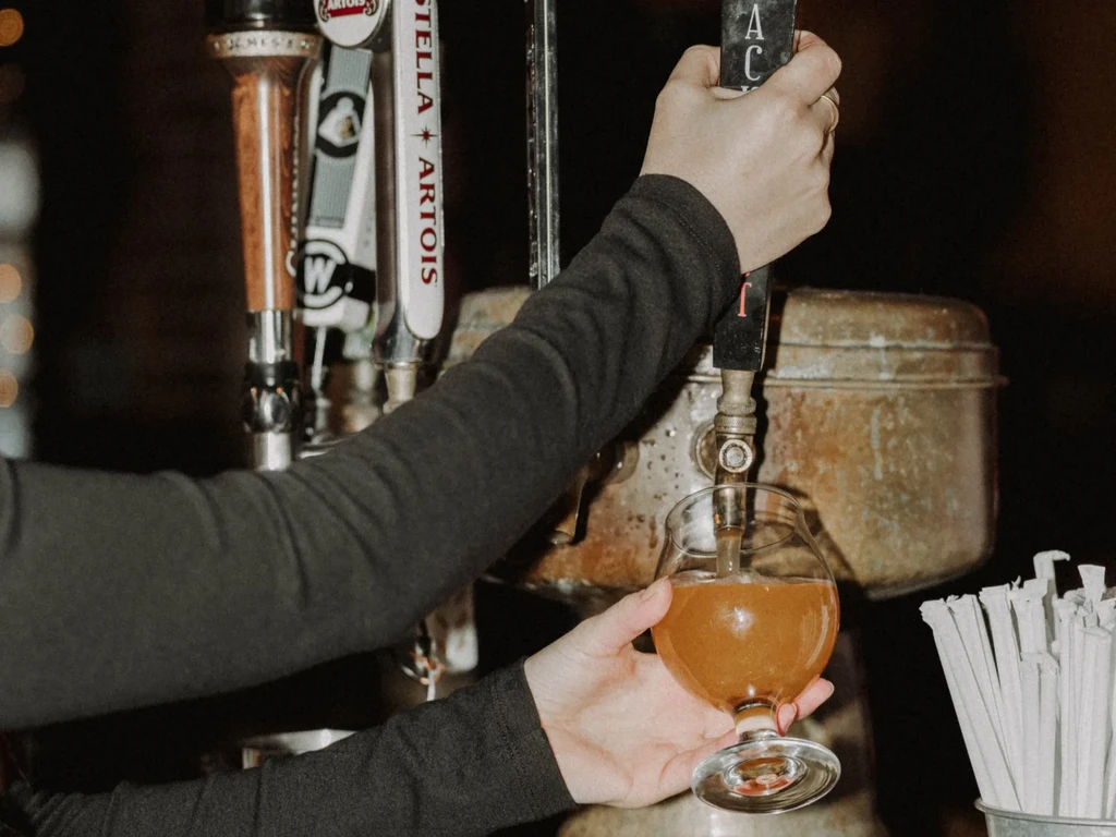 Person's hands pour hazy amber beer from a tap into a stemmed glass.